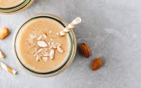 Ingredients for a chocolate almond shake including a glass of almond milk, cocoa powder, and a scoop of chocolate protein powder on a kitchen counter.